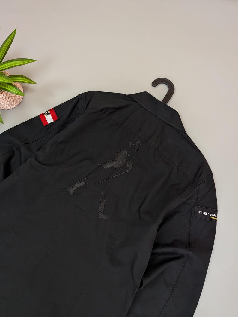 Puede incluir: Camisa negra con botones con un peque&ntilde;o parche de la bandera austriaca en la manga. La parte trasera presenta una gran figura negra estilizada. Una peque&ntilde;a etiqueta en la manga dice "KEEP WAL". La camisa est&aacute; colgada en una percha negra.