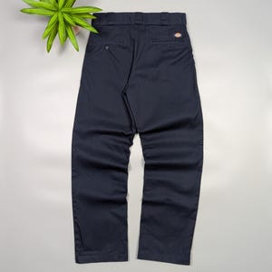 May include: Navy blue work pants with a button closure and back pockets. The pants have a small logo patch on the back pocket. The pants are made of a durable material and are suitable for work or casual wear.