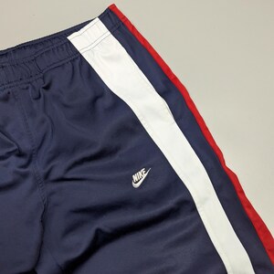 May include: Navy blue tracksuit bottoms with a white stripe and a red accent. The Nike logo is visible. The trousers have an elasticated waistband. The material appears to be a smooth, synthetic fabric.