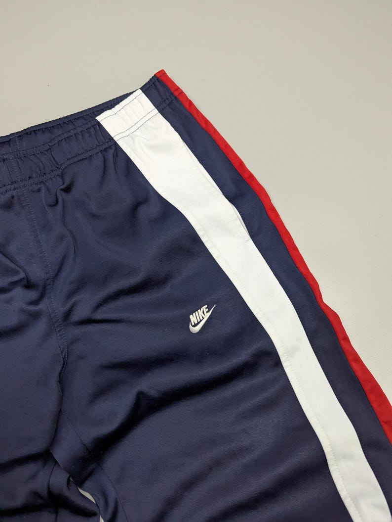May include: Navy blue tracksuit bottoms with a white stripe and a red accent. The Nike logo is visible. The trousers have an elasticated waistband. The material appears to be a smooth, synthetic fabric.