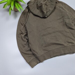 Puede incluir: Una sudadera con capucha verde oliva descolorida con capucha con cord&oacute;n. La parte trasera de la sudadera con capucha es visible, mostrando la capucha y la parte superior de la espalda de la prenda. La tela parece ser un material suave y desgastado. La sudadera es de estilo casual.