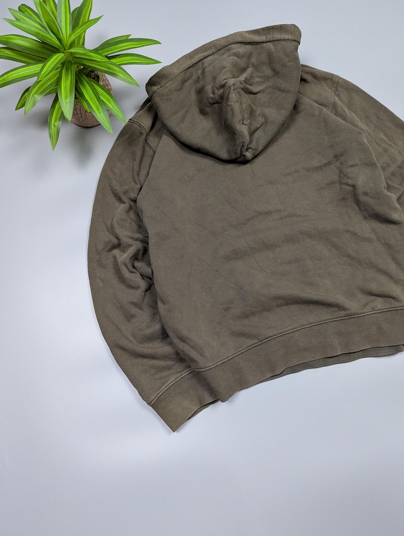Puede incluir: Una sudadera con capucha verde oliva descolorida con capucha con cord&oacute;n. La parte trasera de la sudadera con capucha es visible, mostrando la capucha y la parte superior de la espalda de la prenda. La tela parece ser un material suave y desgastado. La sudadera es de estilo casual.