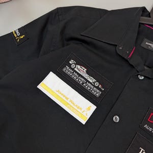 Puede incluir: Camisa negra con botones con m&uacute;ltiples parches bordados. Los parches incluyen el texto "JOHNNIE WALKER RACING" y "TEAM MCLAREN MERCEDES CORPORATE PARTNER". La camisa tiene cuello y pu&ntilde;os abotonados. La camisa est&aacute; colgada en una percha.
