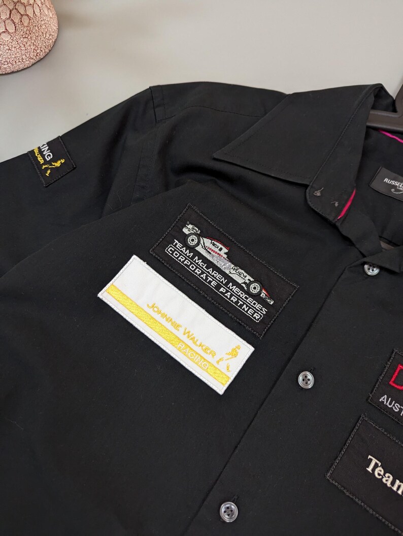 Puede incluir: Camisa negra con botones con m&uacute;ltiples parches bordados. Los parches incluyen el texto "JOHNNIE WALKER RACING" y "TEAM MCLAREN MERCEDES CORPORATE PARTNER". La camisa tiene cuello y pu&ntilde;os abotonados. La camisa est&aacute; colgada en una percha.
