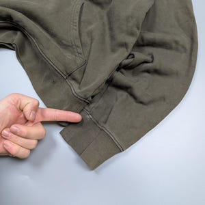 Puede incluir: Sudadera con capucha verde oliva con bolsillo de canguro y pu&ntilde;os acanalados. La prenda est&aacute; hecha de un material suave y desgastado. La imagen muestra un primer plano de la manga y el bolsillo.