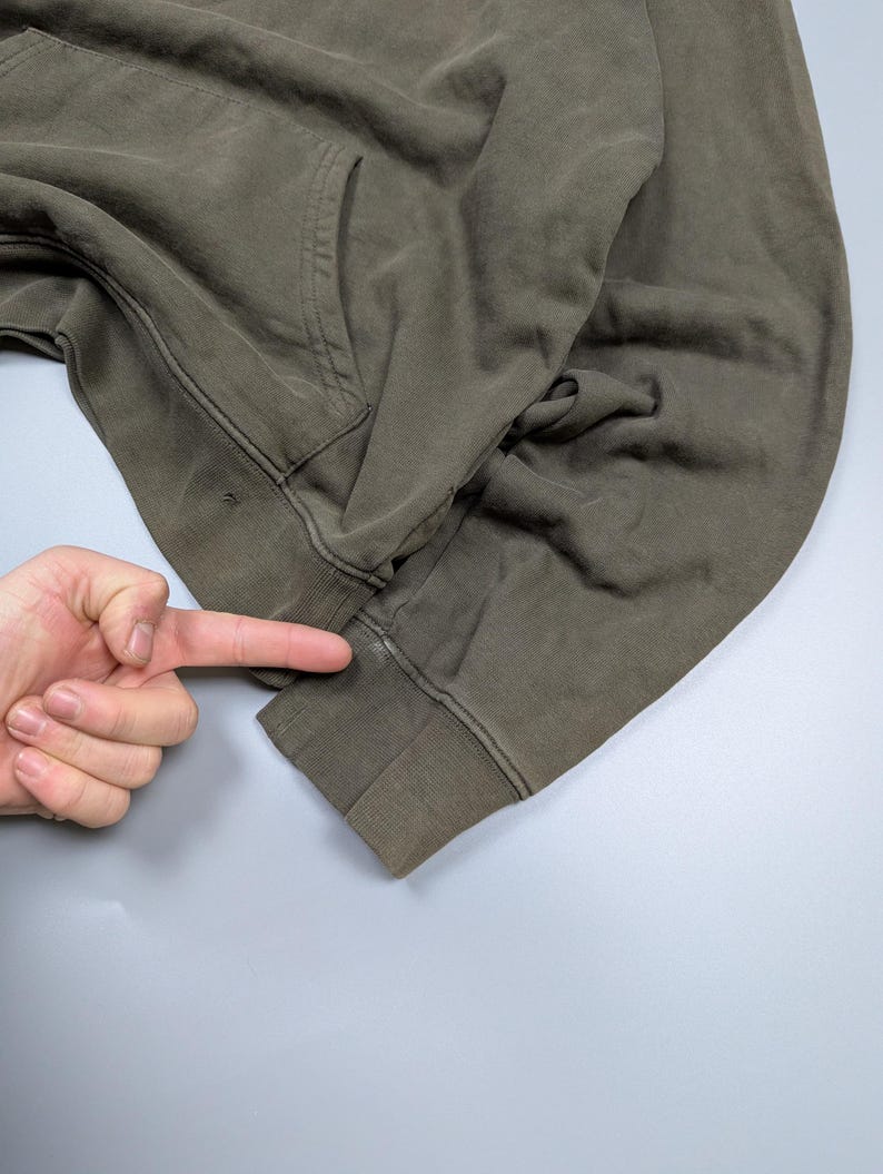 Puede incluir: Sudadera con capucha verde oliva con bolsillo de canguro y pu&ntilde;os acanalados. La prenda est&aacute; hecha de un material suave y desgastado. La imagen muestra un primer plano de la manga y el bolsillo.