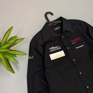 Puede incluir: Camisa negra abotonada con cuello, colgada de una percha negra. La camisa tiene varios parches, incluyendo "DIAGEO Austria" y "Teamplayer" en texto rojo. Tambi&eacute;n es visible un peque&ntilde;o parche de "Johnnie Walker".