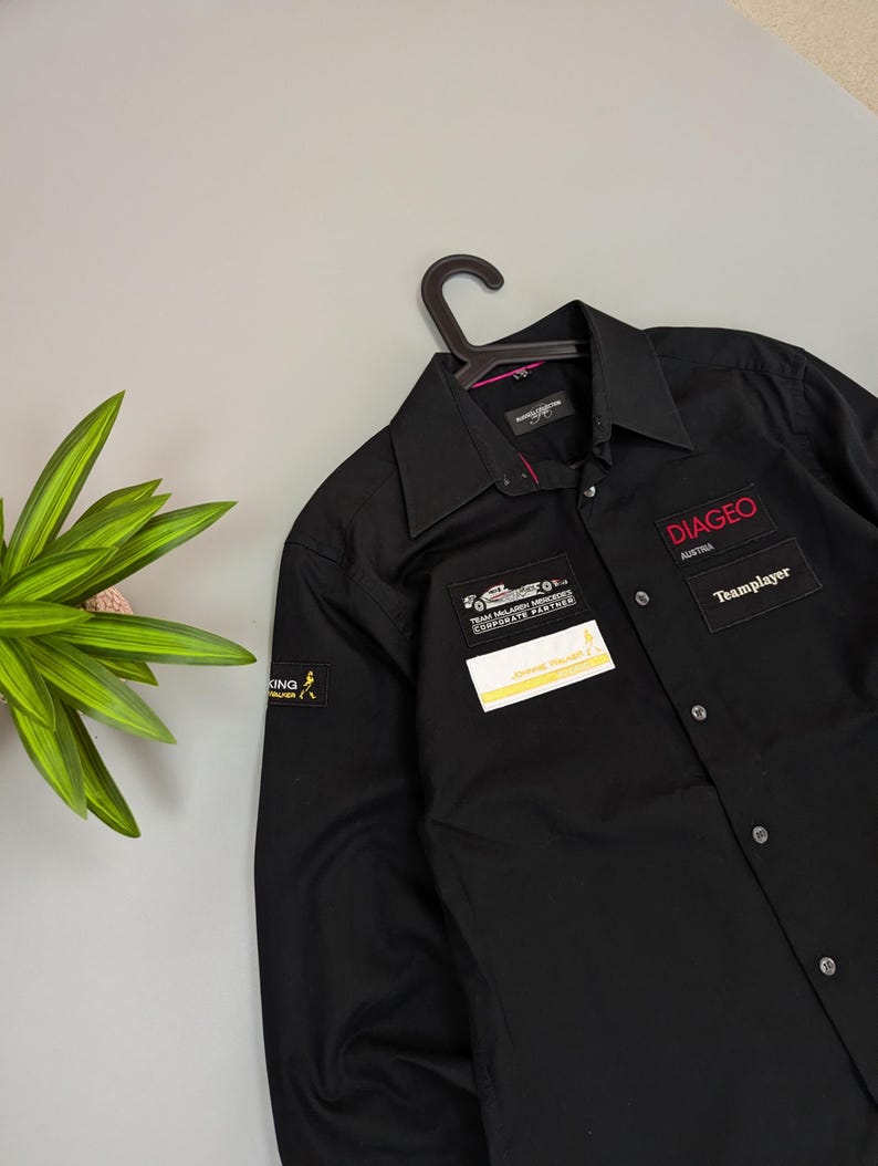Puede incluir: Camisa negra abotonada con cuello, colgada de una percha negra. La camisa tiene varios parches, incluyendo "DIAGEO Austria" y "Teamplayer" en texto rojo. Tambi&eacute;n es visible un peque&ntilde;o parche de "Johnnie Walker".
