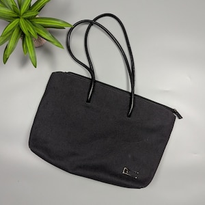 May include: Black tote bag with glossy black handles and a zipper closure. The bag has a rectangular shape and a small gold-colored accent on the front. A green plant is visible in the upper left corner.