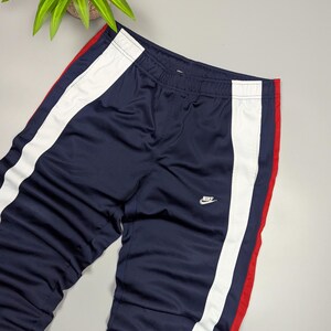 May include: Navy blue track pants with white and red stripes down the sides. The pants have an elasticated waistband and the Nike logo in white. The trousers are made of a shiny, athletic material.