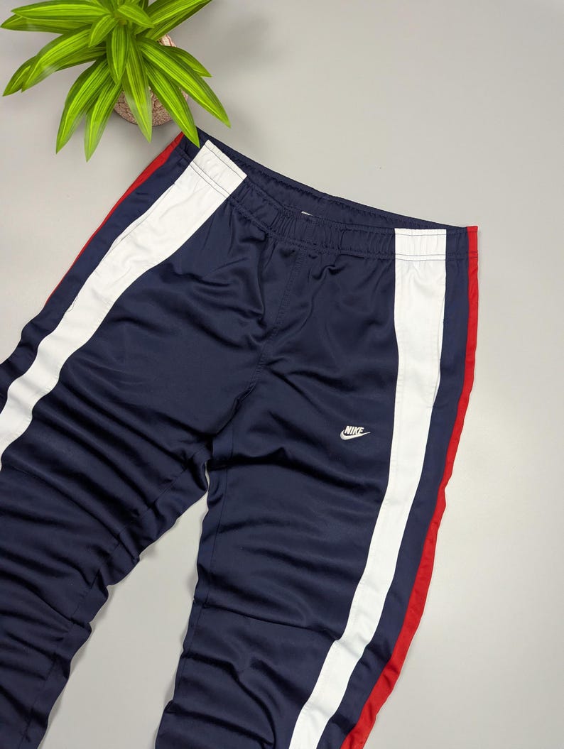 May include: Navy blue track pants with white and red stripes down the sides. The pants have an elasticated waistband and the Nike logo in white. The trousers are made of a shiny, athletic material.