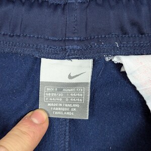 May include: Close-up of navy blue athletic trousers with a tag. The tag displays the Nike logo and size information: SIZE S, HEIGHT 173, GB 28/30, F 44/48, D 44/45. The tag also indicates the trousers were made in Thailand.