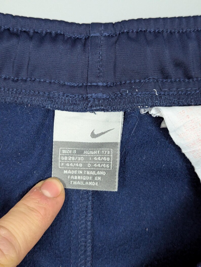May include: Close-up of navy blue athletic trousers with a tag. The tag displays the Nike logo and size information: SIZE S, HEIGHT 173, GB 28/30, F 44/48, D 44/45. The tag also indicates the trousers were made in Thailand.