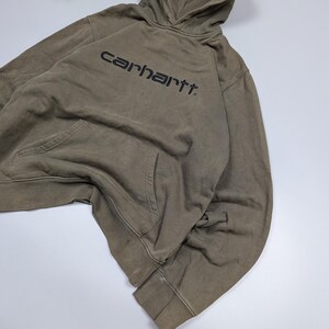 Puede incluir: Una sudadera con capucha Carhartt verde oliva con capucha con cord&oacute;n y bolsillo tipo canguro. El nombre de la marca est&aacute; impreso en negro en el pecho. Los pu&ntilde;os y el dobladillo son acanalados.