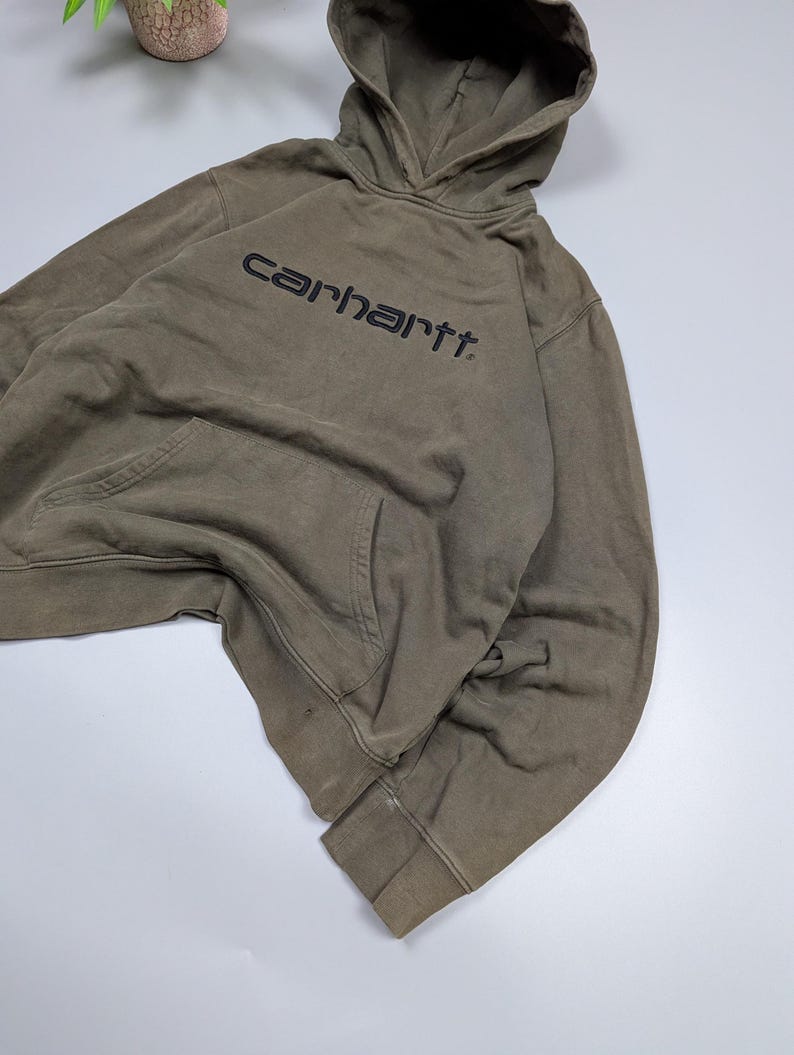 Puede incluir: Una sudadera con capucha Carhartt verde oliva con capucha con cord&oacute;n y bolsillo tipo canguro. El nombre de la marca est&aacute; impreso en negro en el pecho. Los pu&ntilde;os y el dobladillo son acanalados.