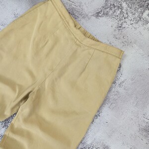 May include: A pair of light tan trousers featuring a flat front waistband with a subtle elasticized back section, vertical seam detailing running down the front, and a clean, solid-colored fabric construction with a smooth, matte finish.