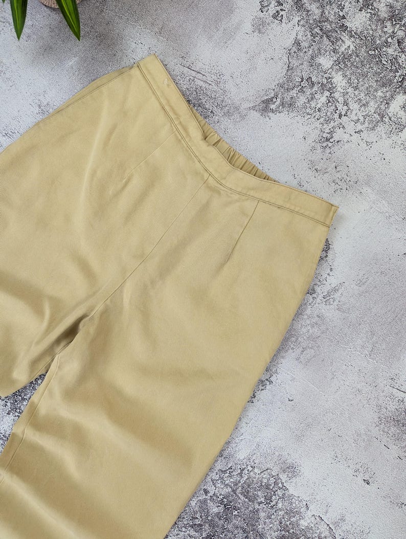 May include: A pair of light tan trousers featuring a flat front waistband with a subtle elasticized back section, vertical seam detailing running down the front, and a clean, solid-colored fabric construction with a smooth, matte finish.