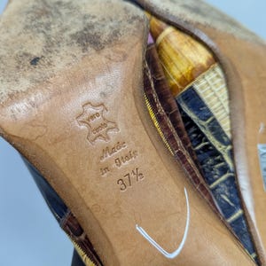 May include: Close-up of the soles of a pair of high-heeled shoes. The soles are brown leather, with the text "Made in Italy" and size "37 1/2" embossed. The shoes have a reptile-like pattern in shades of brown, yellow, and blue.