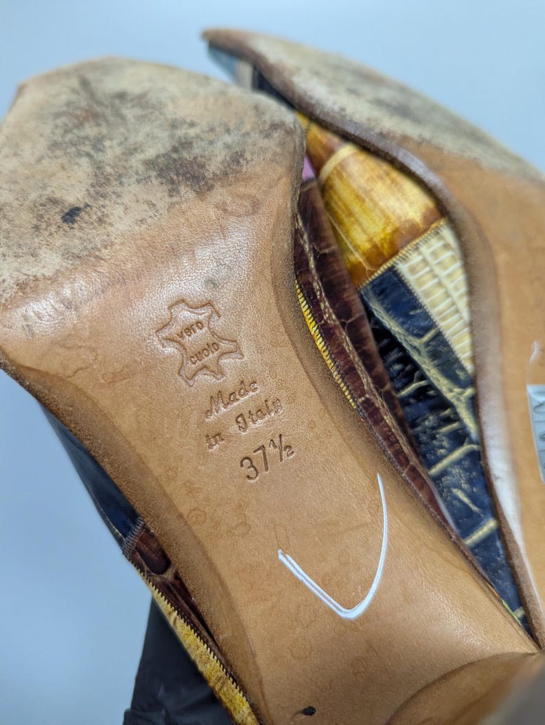 May include: Close-up of the soles of a pair of high-heeled shoes. The soles are brown leather, with the text "Made in Italy" and size "37 1/2" embossed. The shoes have a reptile-like pattern in shades of brown, yellow, and blue.