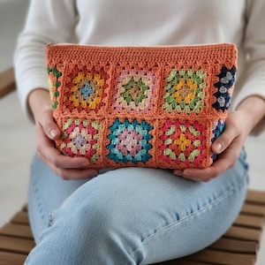 May include: A crochet pouch in a warm orange colour. The front features a patchwork design of colourful granny squares in shades of pink, blue, green, and yellow. The pouch has a zip closure and is held in someone's hands.