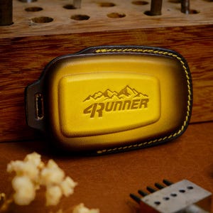 May include: A yellow and black key fob cover featuring a mountain graphic and the text 4RUNNER embossed on the center. The cover has a smooth texture, a rectangular shape with rounded corners, and is stitched with yellow thread along the edges.