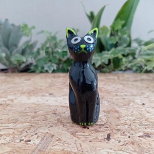 Könnte beinhalten: Glänzende schwarze Keramik-Katzenfigur mit grünen und weißen Akzenten. Die Katze hat große Augen, eine blau gepunktete Nase und grün gefärbte Ohren und Pfoten. Die Figur sitzt aufrecht.