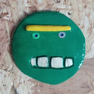 Può includere: Un volto rotondo e verde, in stile cartone animato, con un sopracciglio giallo, due piccoli occhi rotondi e un ampio sorriso con denti. Il volto ha una finitura lucida ed è posto su uno sfondo strutturato marrone chiaro. Il design è stravagante e giocoso.