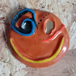 May include: A ceramic smiley face sculpture with an orange base color. The sculpture features a black and blue eye, a red line, a yellow smile, and black dots. The sculpture is on a light brown surface.