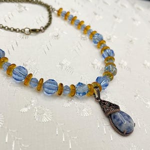 May include: A beaded necklace featuring a mix of light blue and amber-colored beads. The necklace has a bronze-colored chain and a teardrop-shaped pendant with a blue and white stone.