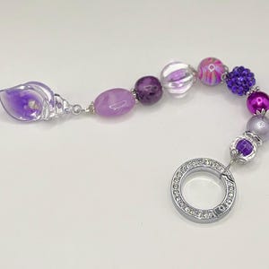 May include: A beaded charm bracelet featuring a variety of purple beads, including a clear, shell-shaped bead with purple accents. The bracelet has a silver clasp with clear rhinestones.