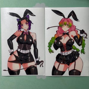 May include: Two anime-style stickers featuring female characters in black bunny outfits. One has purple hair and the other has pink hair. Both are holding whips. The stickers have a white background.