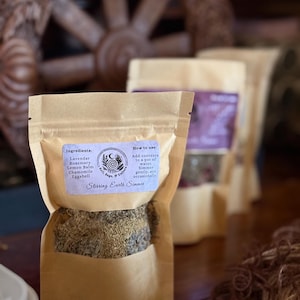 May include: Close-up of several kraft paper pouches filled with dried herbs. The front pouch has a label that reads "Stirring Earth Simmer" and lists ingredients like lavender and rosemary. Instructions are also included on the label.