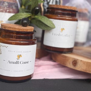 May include: A selection of amber glass jar candles with cork lids. The labels read "Amalfi Coast" and "Fresh Coffee." The candles are presented with greenery and a wooden board. A price tag is visible in the background.