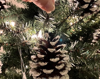 Puede incluir: Primer plano de un árbol de Navidad decorado con adornos de piña. Las piñas están cubiertas de purpurina blanca y cuelgan de las ramas del árbol. También hay un adorno de carámbano transparente colgando del árbol.