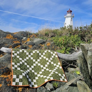 May include: A square quilt featuring a repeating geometric pattern of olive green and white fabric squares. The quilt is finished with a solid orange fabric binding along the edges and displays a visible grid of white stitching across the surface.