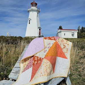 Peut inclure: Une courtepointe en patchwork avec un motif en étoile composé de panneaux de tissu dans des tons orange, blanc, crème et violet clair. La courtepointe présente une bordure jaune et est drapée sur une chaise en bois usé devant un phare blanc.