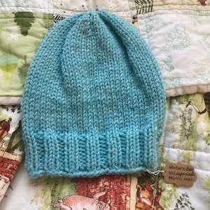 May include: A light blue knitted baby hat made of merino wool. The hat has a ribbed brim and a simple, classic design. A small tag attached to the hat reads "Newborn-Baby 100% Superwash Merino wool". The hat is displayed on a patterned quilt.
