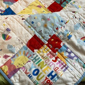 May include: A patchwork quilt featuring colorful squares with patterns of rainbows, clouds, suns, and kites. The fabric includes text that reads "pure magic," "shine," "you are," and "happy." The quilt is finished with a light blue border.