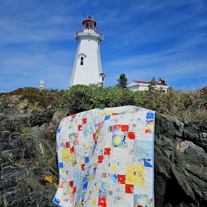 May include: A patchwork quilt featuring a variety of colorful squares with illustrations of suns, rainbows, and clouds, finished with a light blue border. The quilt is draped over dark, jagged rocks in front of a white lighthouse under a blue sky.