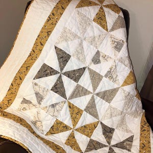 以下が含まれることがあります： 茶色と金の幾何学模様が施された白いキルト。キルトは、花柄の茶色と金の生地の縁取りがされています。