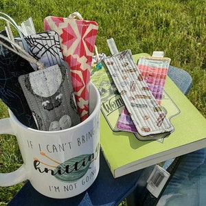 May include: A white ceramic mug with the text "If I can't bring my knitting I'm not going" and a depiction of knitting needles and yarn. The mug is filled with colorful fabric bookmarks.