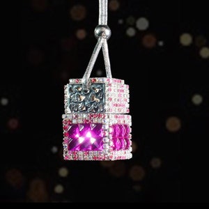 May include: A pink and silver car air freshener with a crystal-like design. The air freshener is hanging from a silver chain.