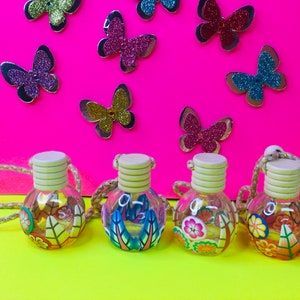 May include: Four small glass bottles with colorful floral and leaf designs and wooden stoppers. The bottles are hanging from a string and are on a yellow surface. The background is a bright pink surface with silver and colorful butterflies.