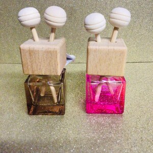 May include: Two car air fresheners with wooden tops and white ball diffusers. The air fresheners are filled with pink and brown liquid.