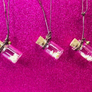May include: Three small glass bottles with cork tops and brown cords hanging from them. Each bottle contains white glitter and is hanging against a pink sparkly background.