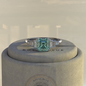 May include: A silver ring with a square-shaped green gemstone surrounded by smaller clear gemstones. The ring is displayed in a box with the words "Royal Blue Lab Diamond Jewelry" printed on the side.
