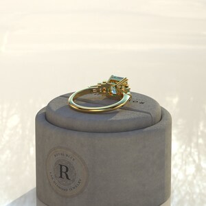 May include: A gold ring with a large emerald-cut aquamarine gemstone surrounded by smaller emerald-cut gemstones. The ring is sitting in a gray box with the words "Royal Blue Diamond Jewelry" printed on the side.