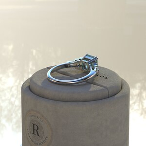 May include: A silver ring with a large emerald-cut green gemstone and two smaller emerald-cut green gemstones on the band. The ring is sitting on a gray box with the words "Royal Blue Lab Diamond Jewelry" printed on the side.
