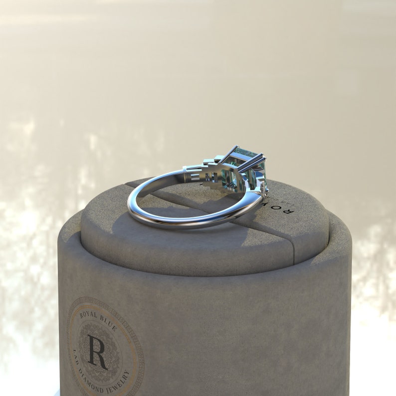 May include: A silver ring with a large emerald-cut green gemstone and two smaller emerald-cut green gemstones on the band. The ring is sitting on a gray box with the words "Royal Blue Lab Diamond Jewelry" printed on the side.