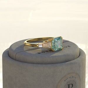May include: A gold ring with a large, emerald-cut green gemstone surrounded by smaller, baguette-cut diamonds. The ring is sitting in a gray box with the words "Royal Blue Lab Grown Diamonds" printed on it.